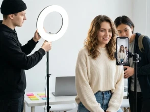 Three people recording a video indoors, with one person on camera and others adjusting a ring light and smartphone.